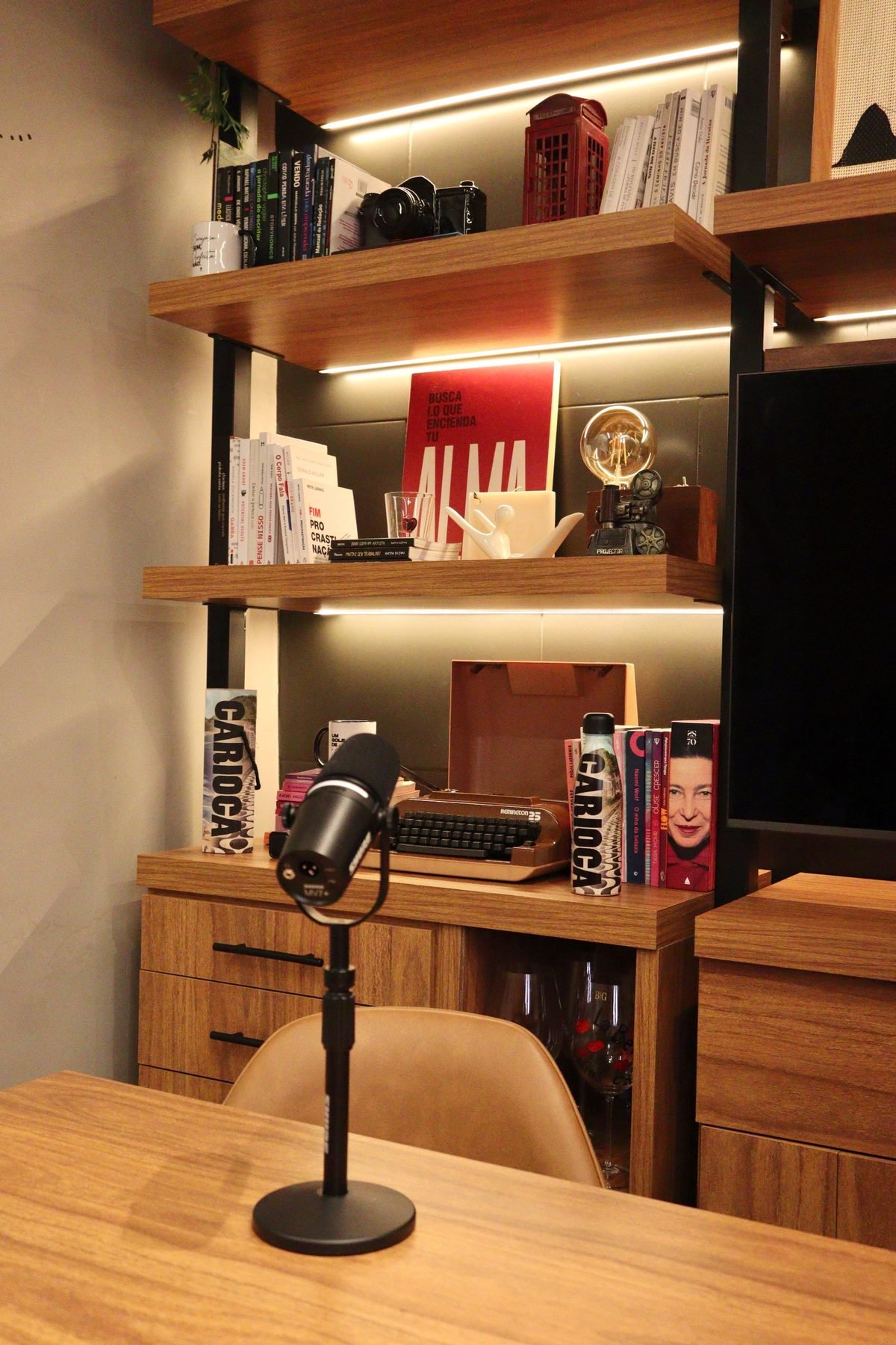 Microfone na mesa de gravação do Carioca Hub, com a estante iluminada ao fundo